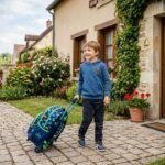 Un jeune garçon sourit en marchant sur un chemin pavé tout en tirant son sac à dos à roulettes à motifs de dinosaures devant une maison en pierre.