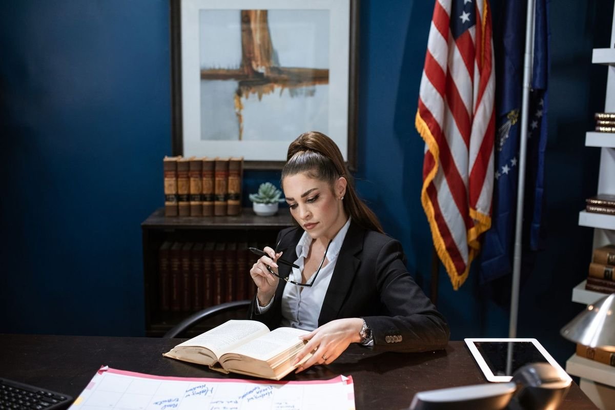 Femme en costume lit un livre à son bureau, tablette et documents à portée de main, drapeau américain et étagères dans le fond Femme en costume lit un livre à son bureau, tablette et documents à portée de main, drapeau américain et étagères dans le fond