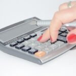 Une main aux ongles rouges appuie sur une calculatrice avec un stylo