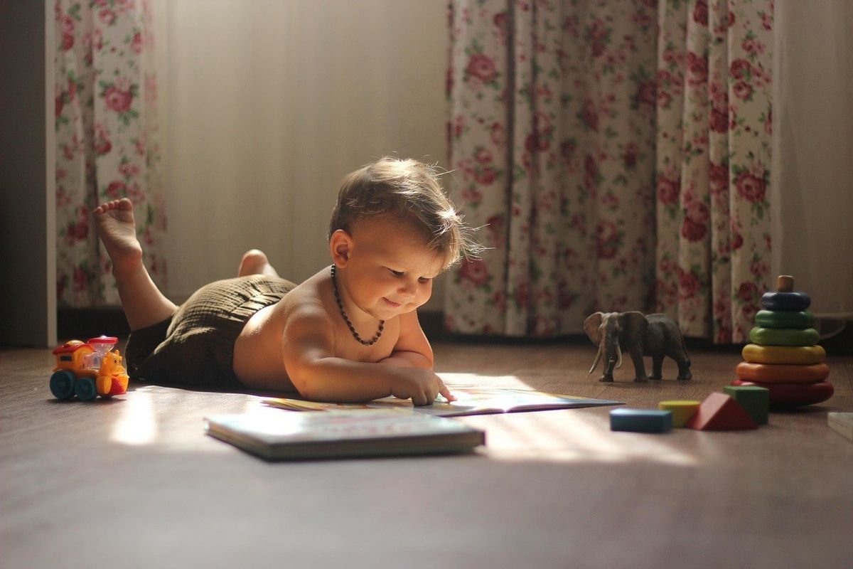 Enfant nu lisant un livre sur le sol, jouets et rideaux floraux à proximité